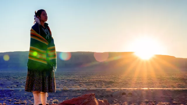 Native American woman wearing traditional clothing overlooks vista with sunset behind her.